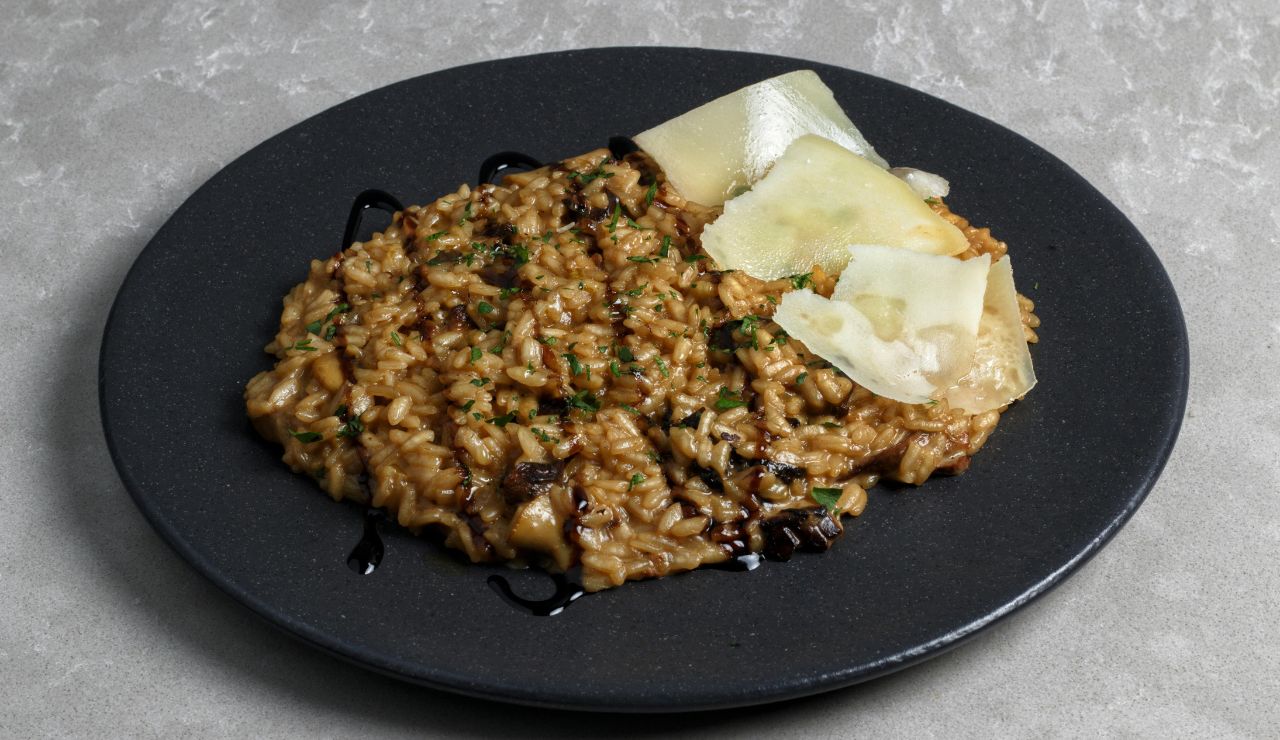 A black plate of mushroom risotto is garnished with fresh herbs, balsamic glaze, and Parmesan shavings.