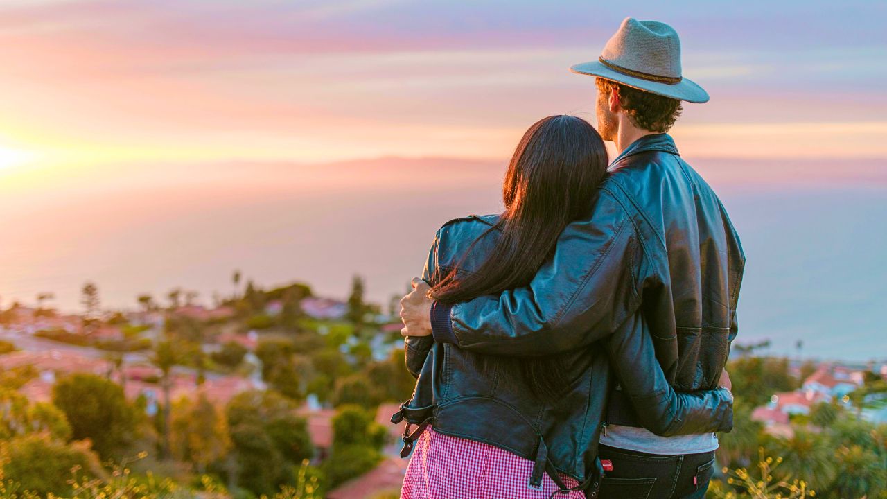 A couple in jackets stands outdoors, facing a sunset over a coastal landscape with houses, trees, and the distant ocean.