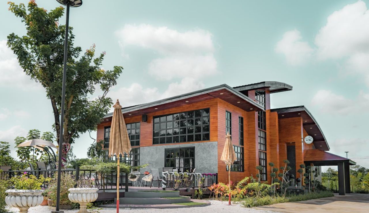 Modern wooden café building with large glass windows, outdoor seating, and umbrellas on a sunny day.