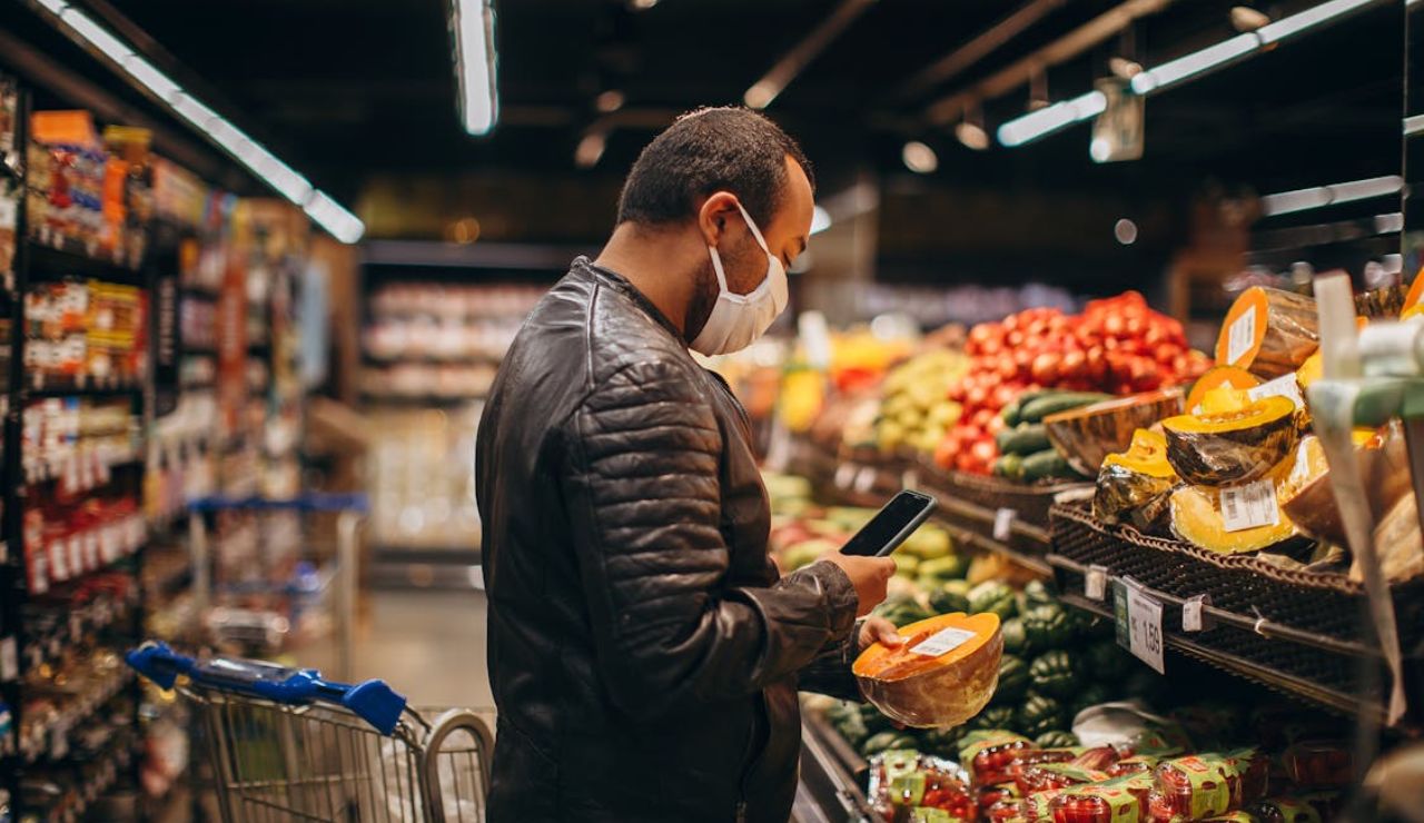 Man wearing a mask shopping for produce in a grocery store while holding a smartphone.