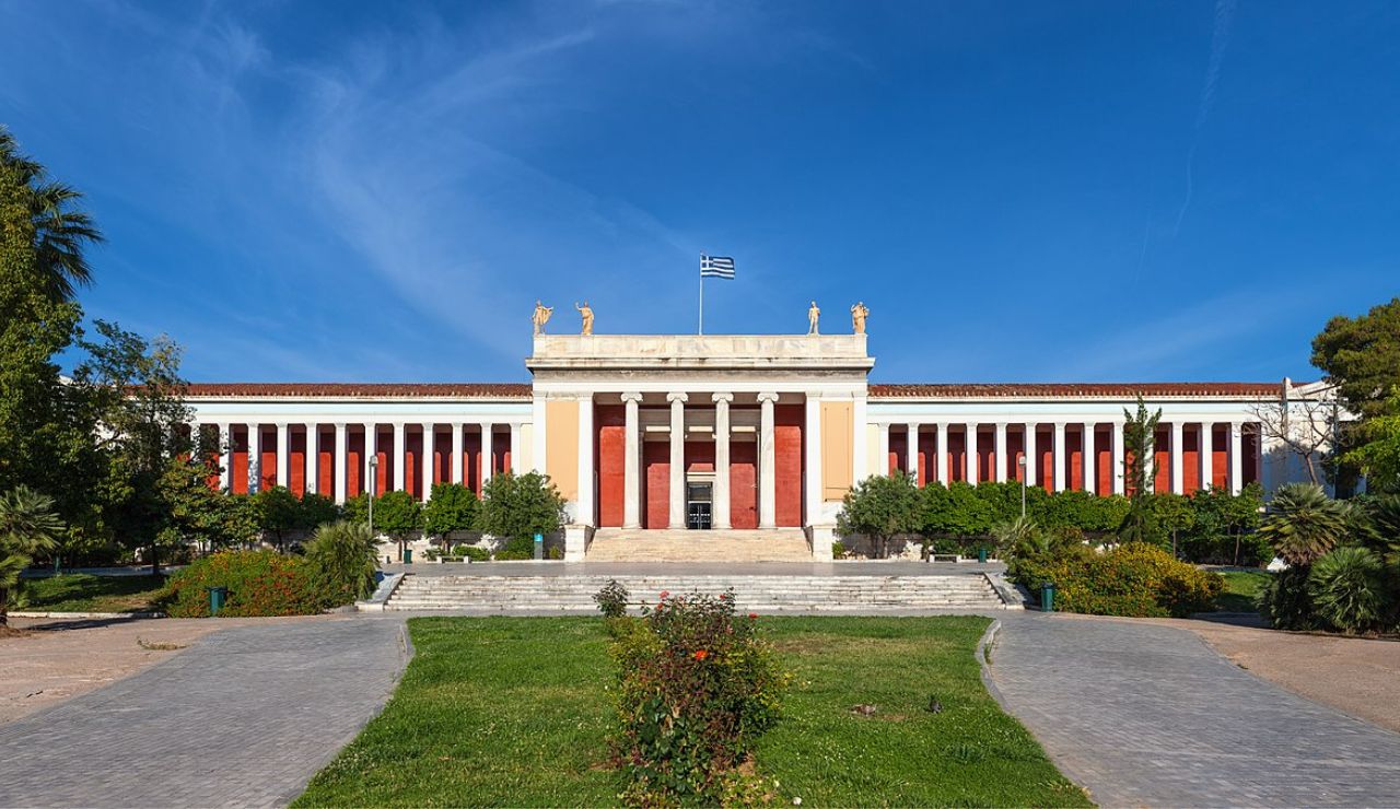 Front view of the National Archaeological Museum of Athens with neoclassical architecture, red columns, and the Greek flag flying above.