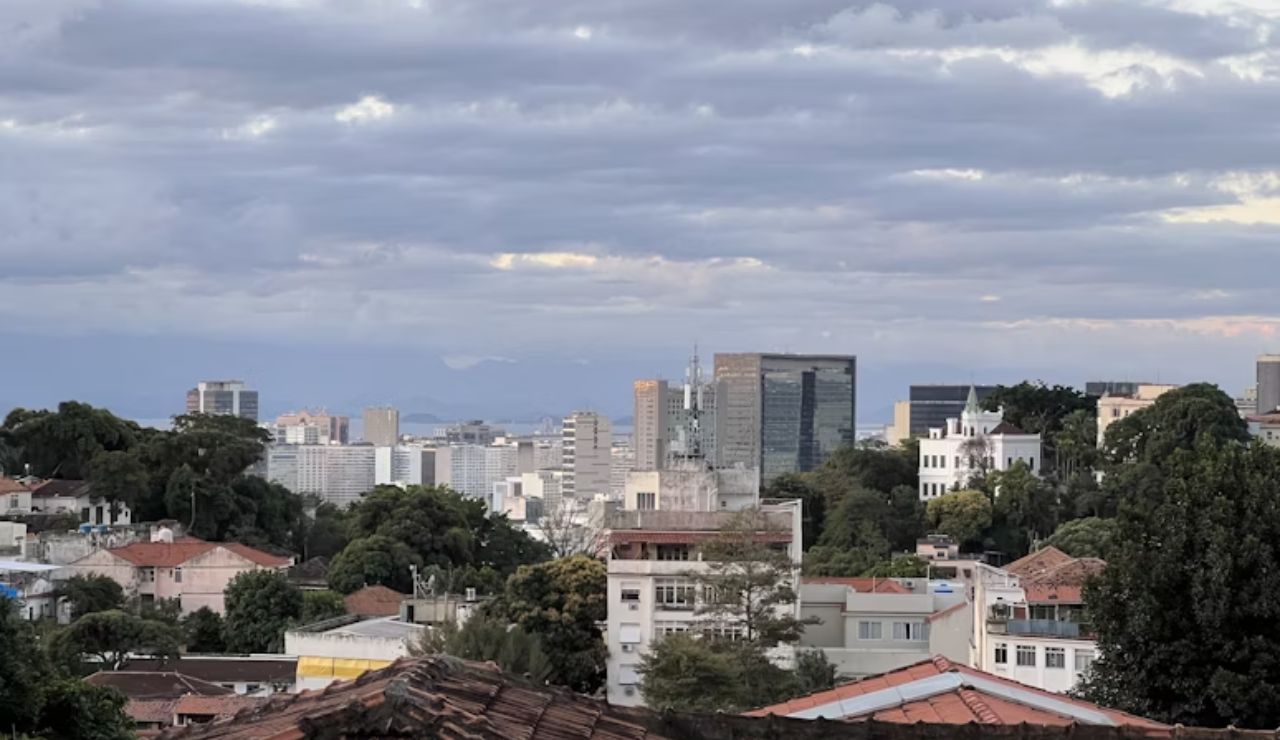 A view of a city skyline with tall buildings, rooftops, and mountains in the distance under a cloudy sky.