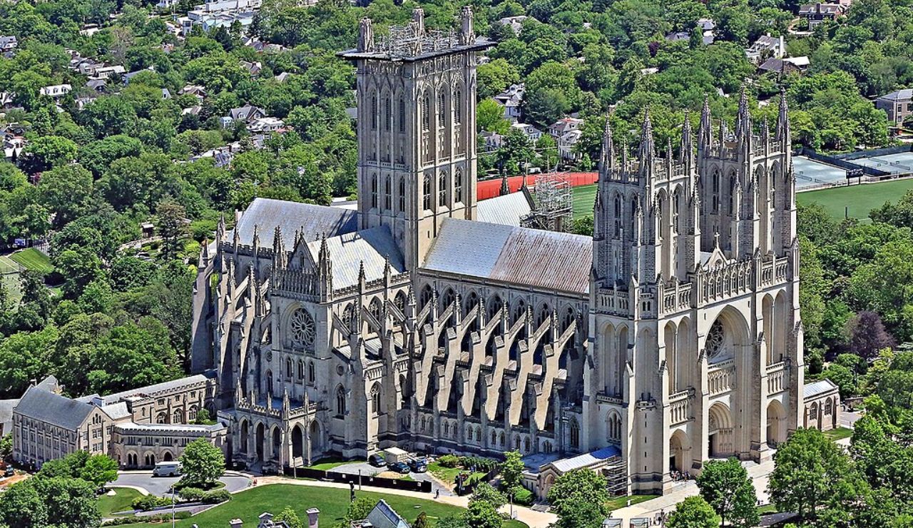 Aerial view of a grand Gothic-style cathedral with towering spires, flying buttresses, and intricate stonework, surrounded by lush greenery and suburban neighborhoods.