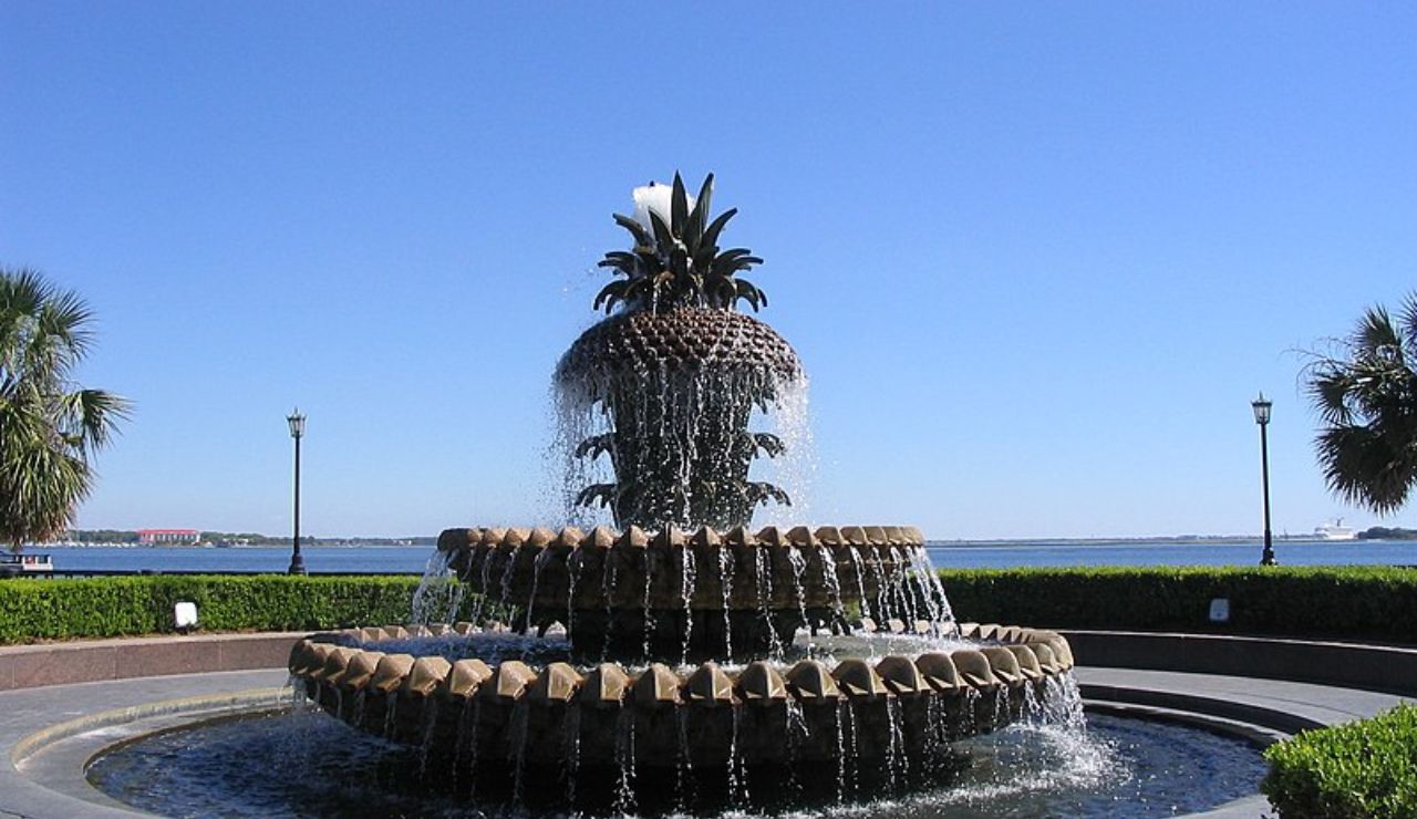 Tiered pineapple-shaped fountain with cascading water, set near a waterfront with palm trees and a clear blue sky.