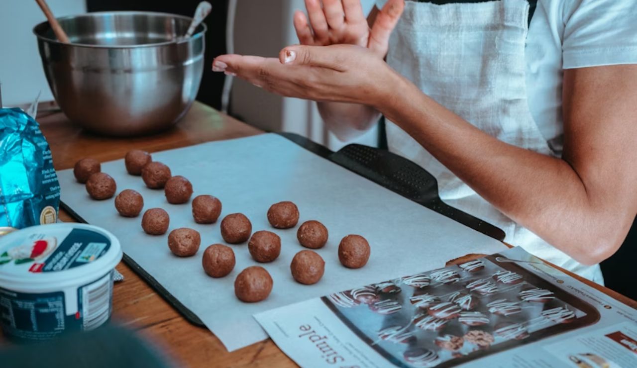 Person shaping chocolate truffles by hand on a parchment-lined tray, with ingredients and a mixing bowl on a wooden table.