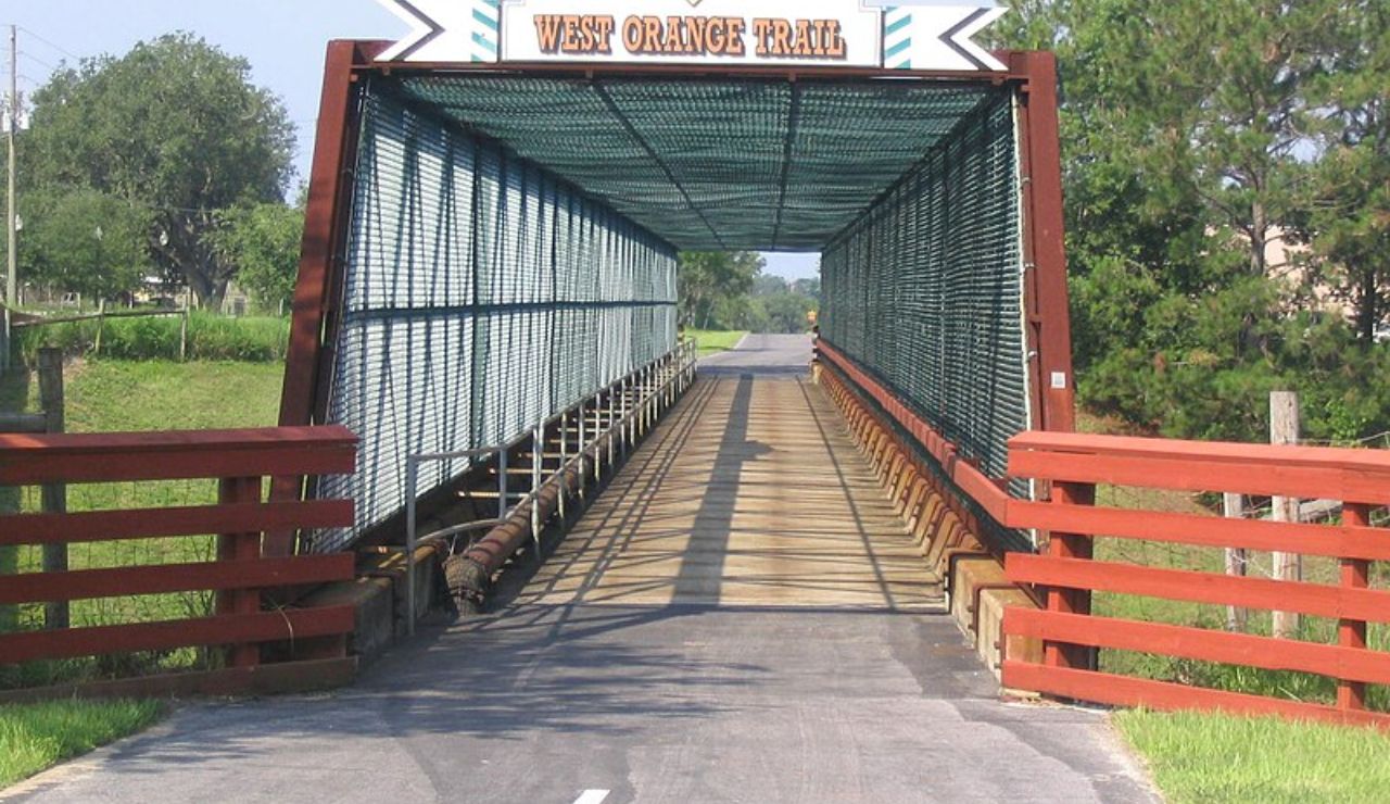 Covered pedestrian and bike bridge labeled "West Orange Trail" with metal fencing and red wooden railings.