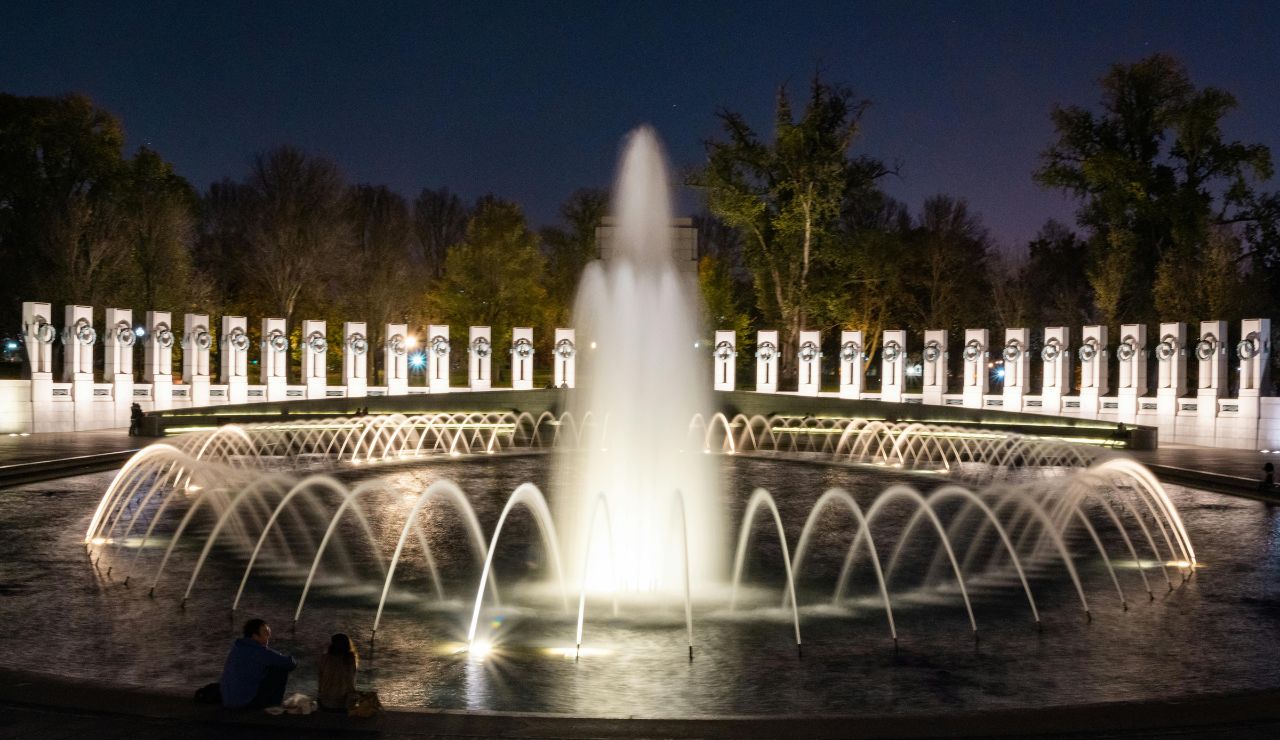 Illuminated World War II Memorial fountain at night, with arched water jets and a circular column backdrop, surrounded by trees and visitors.