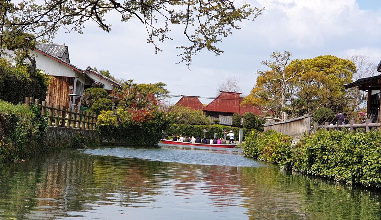 A small boat with passengers floats on a calm canal amid greenery and traditional buildings under a partly cloudy sky.