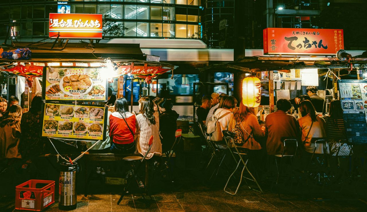 People dine at outdoor food stalls on a busy, lantern-lit Japanese city street at night.