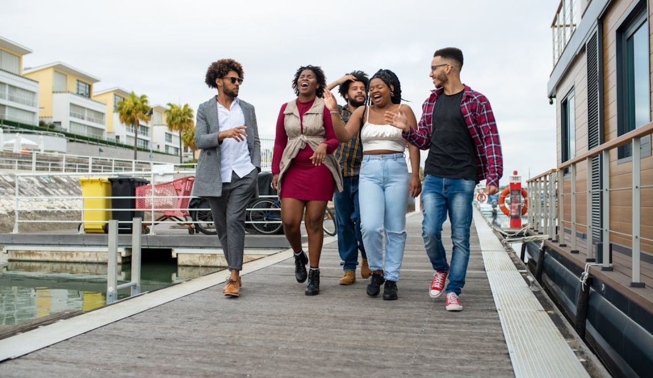 Group of friends laughing and walking on a marina dock near boats and modern buildings.
