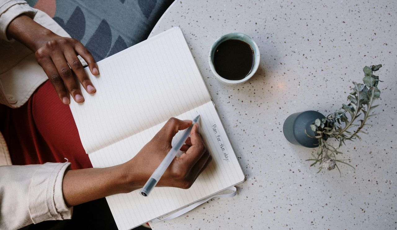 Person writing in a notebook at a table with coffee and a small plant.