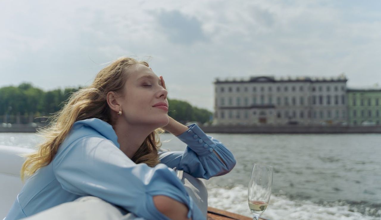Woman in a blue blouse relaxing on a boat during a river cruise, eyes closed and smiling with a glass of champagne beside her and a historic building in the background.