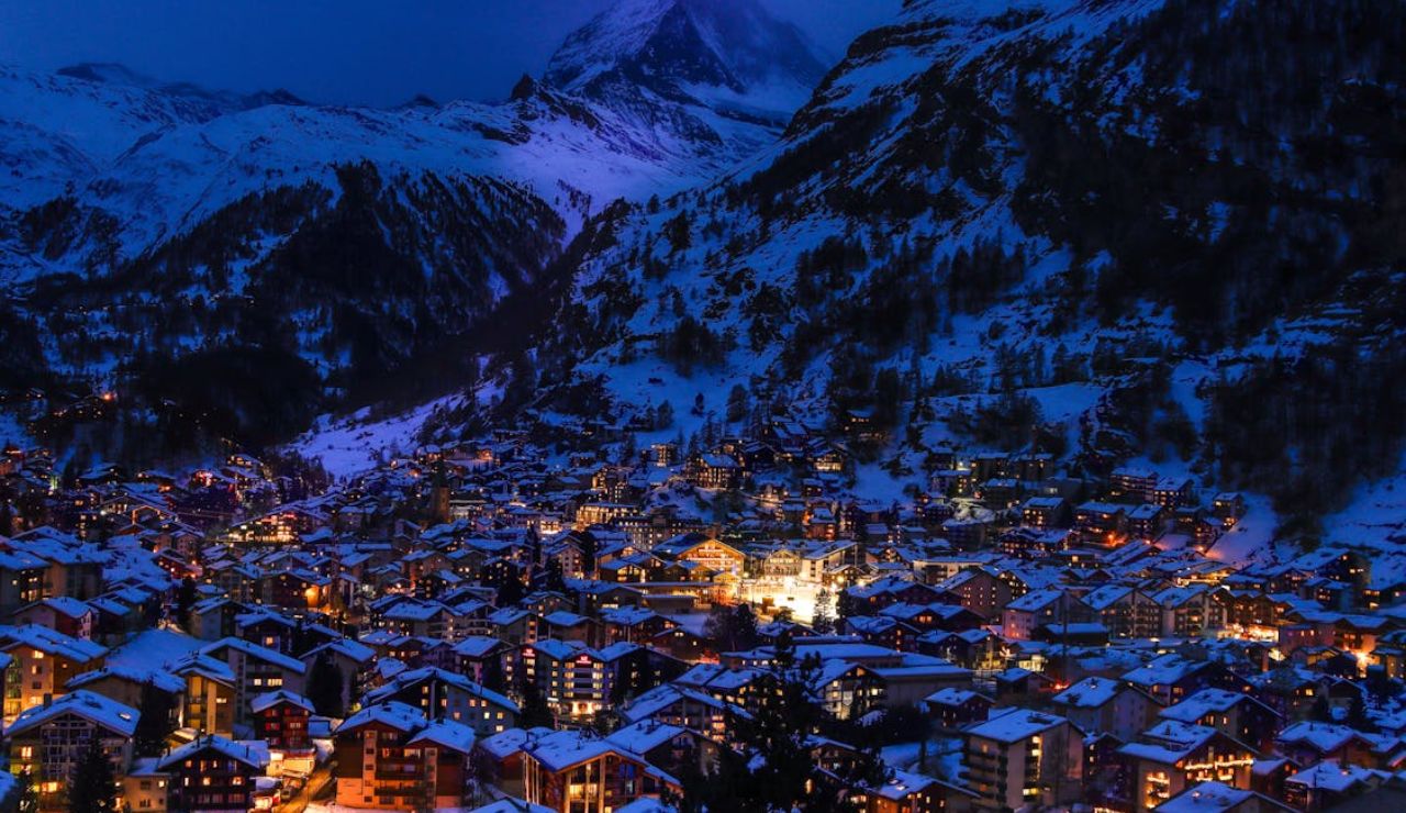Snow-covered alpine village glowing with warm lights at night, nestled in a valley surrounded by towering mountains.