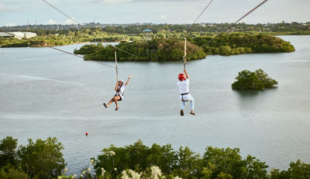 Two people ziplining over a scenic river surrounded by lush greenery.