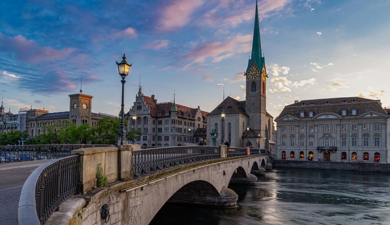 Stone bridge leading to a historic European church with a tall green spire, set against a colorful evening sky over a calm river.
