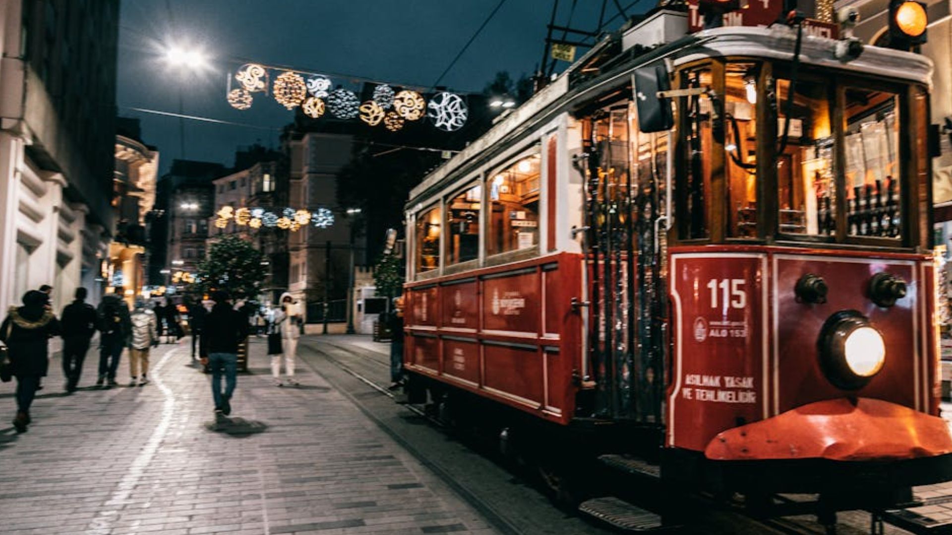 A red tram on a cobblestone street at night, with festive lights and people walking.