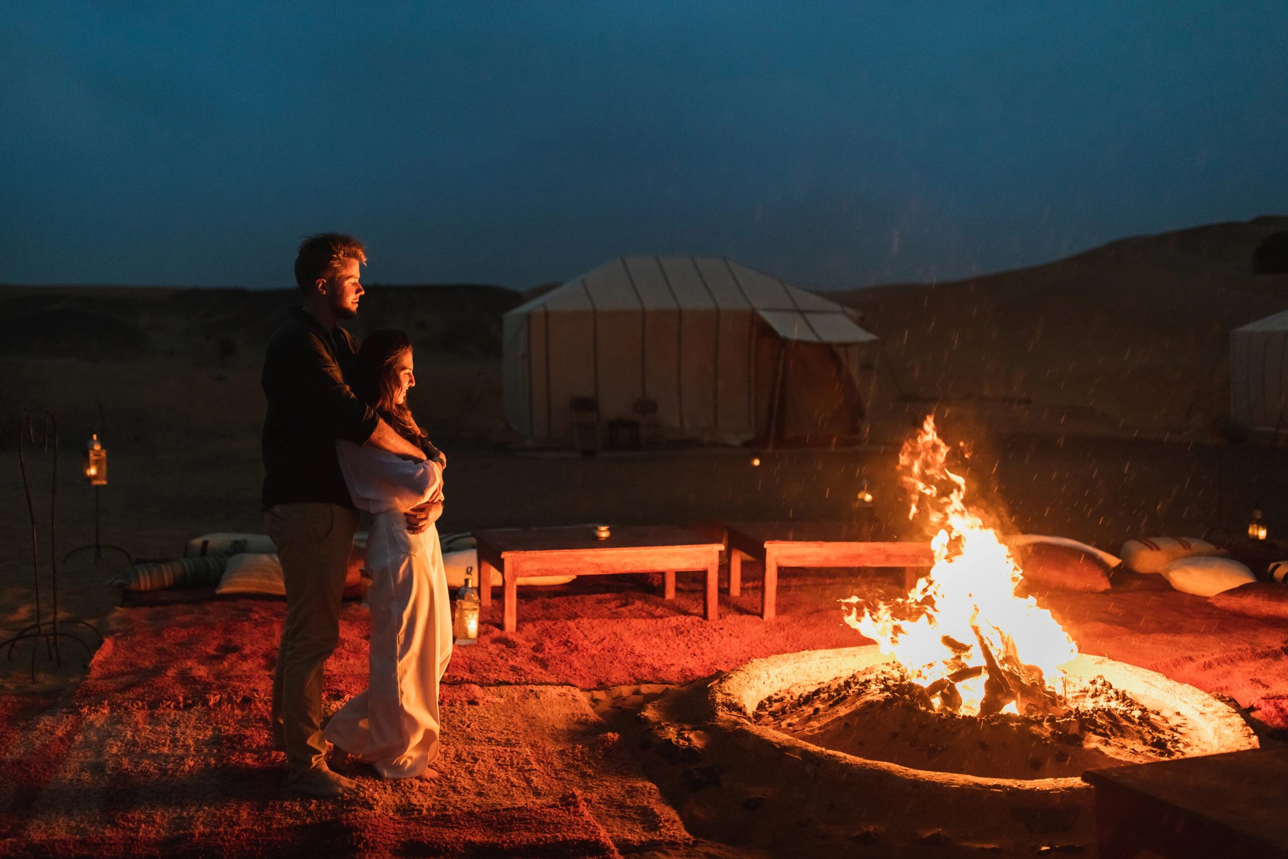 A couple stands on a red carpet by a campfire, with glamping cabins, tents, and benches under a dark evening sky.
