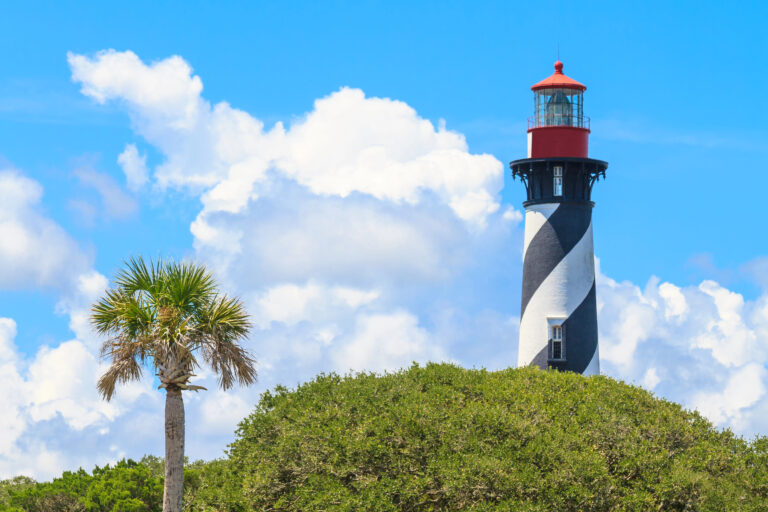 Lighthouse in St. Augustine perched out of tree tops against a blue cloudy sky.