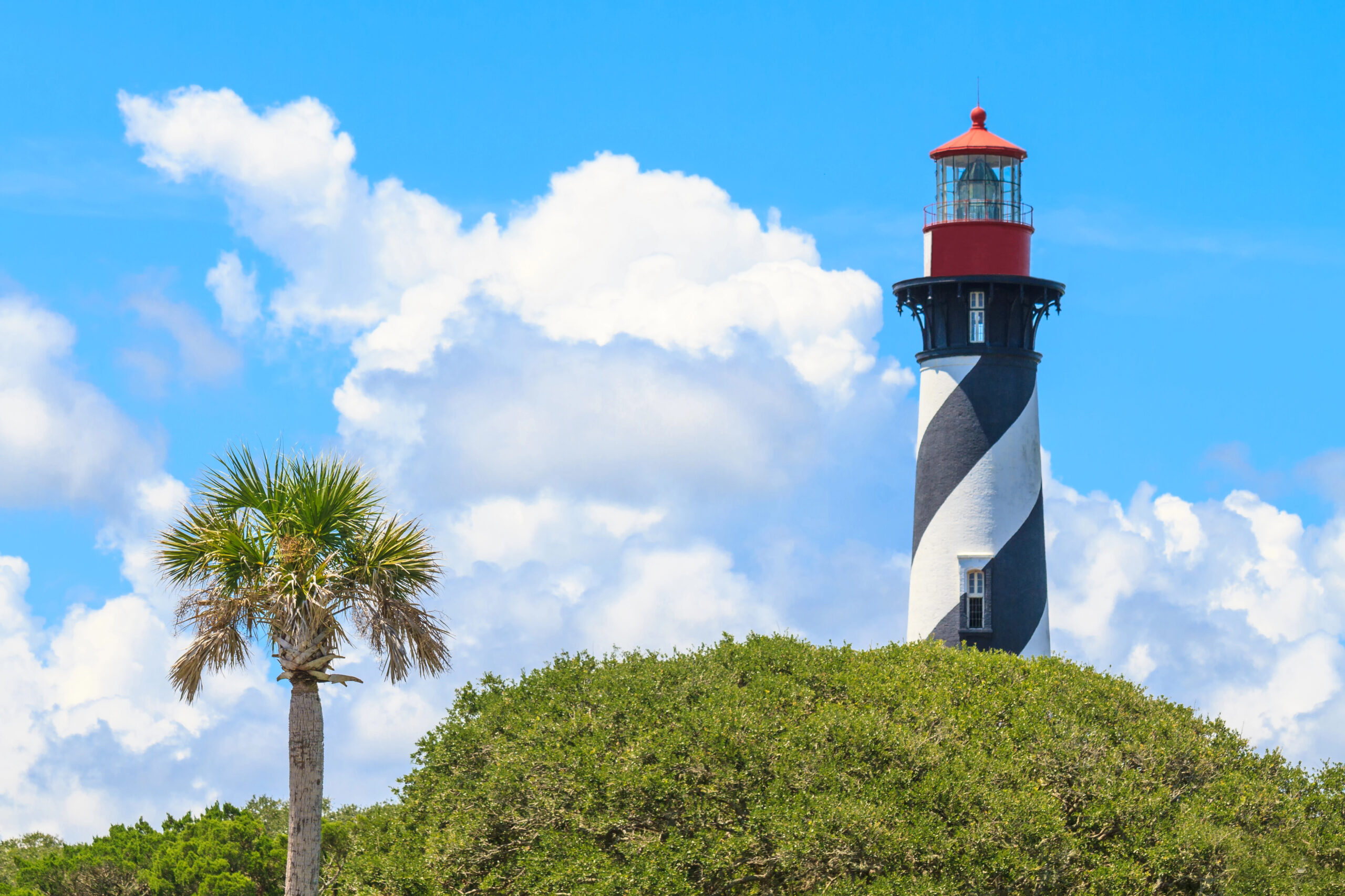 Lighthouse in St. Augustine perched out of tree tops against a blue cloudy sky.