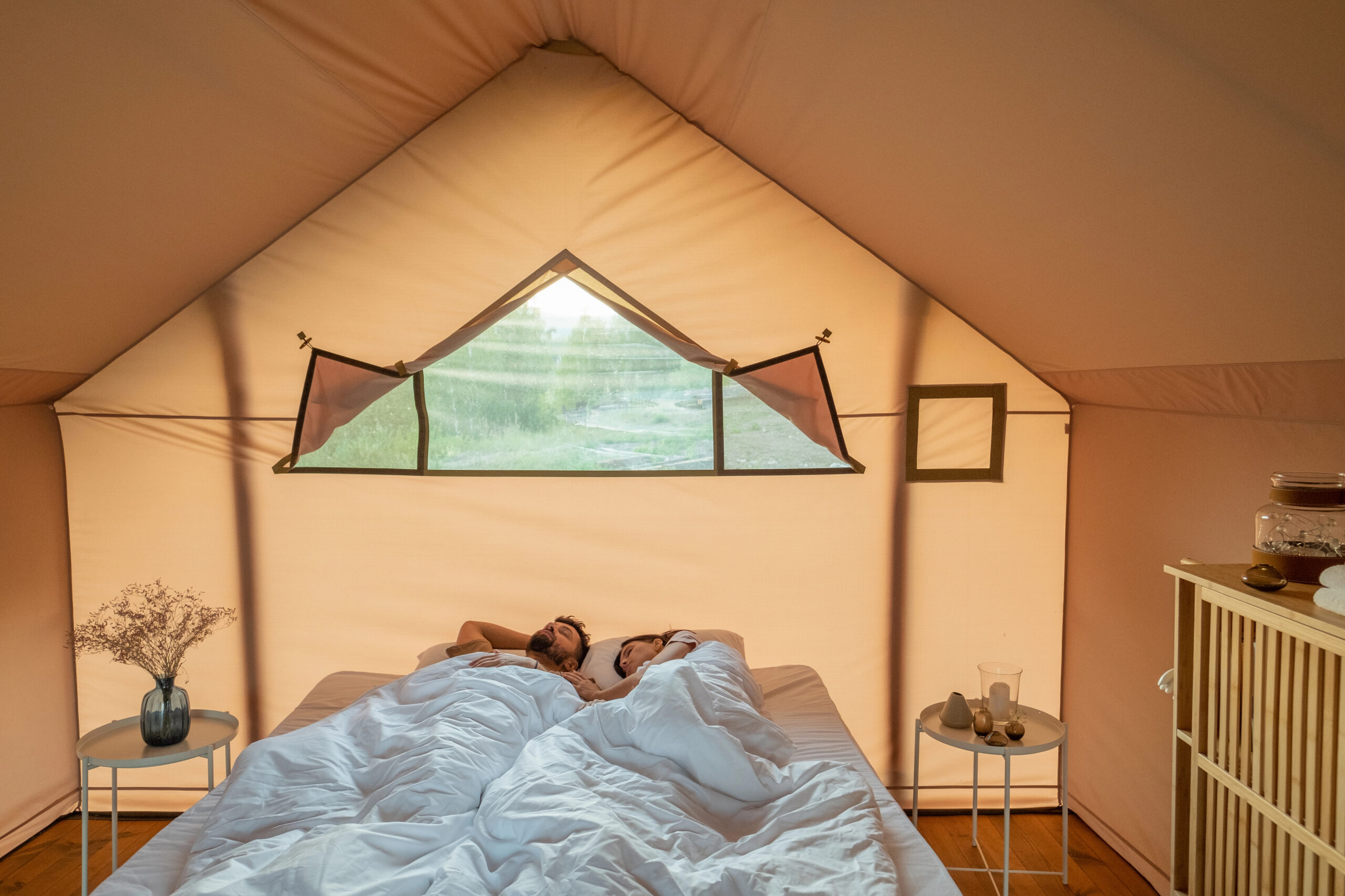 Two people lie under a white blanket in a beige tent with an open window, small tables, a vase, and a cabinet.