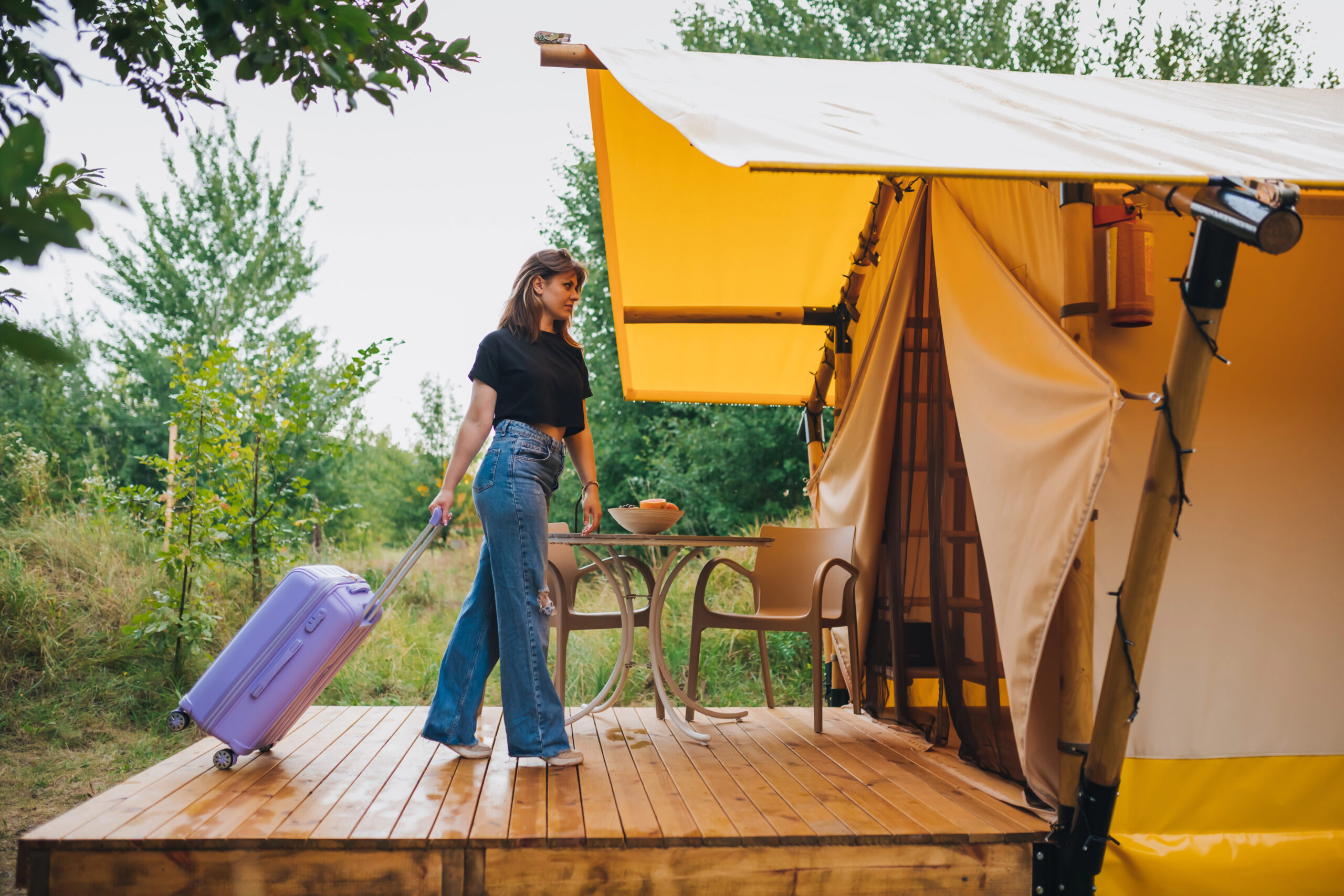 A woman with a purple suitcase stands on a wooden deck near a glamping tent, cabins, and a table with chairs among trees.