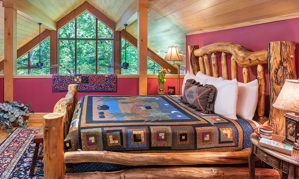 A rustic cabin king bedroom with an A-frame window with views of the forest.