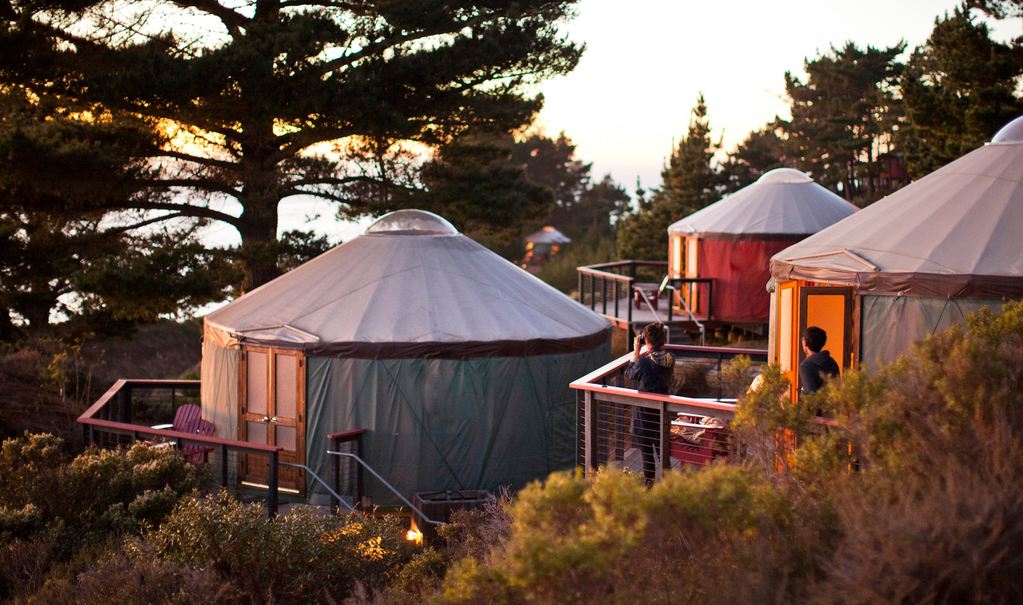 Round canvas glamping cabins with domed roofs sit on a wooded hillside at sunset, people visible on decks and among trees.