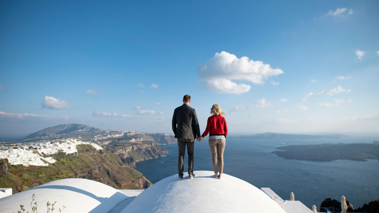 Couple holding hands while standing on a white rooftop overlooking the sea in Santorini.