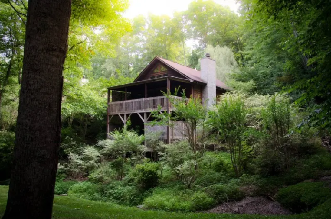 A cabin in a sun-illuminated woods with a balcony and front porch.