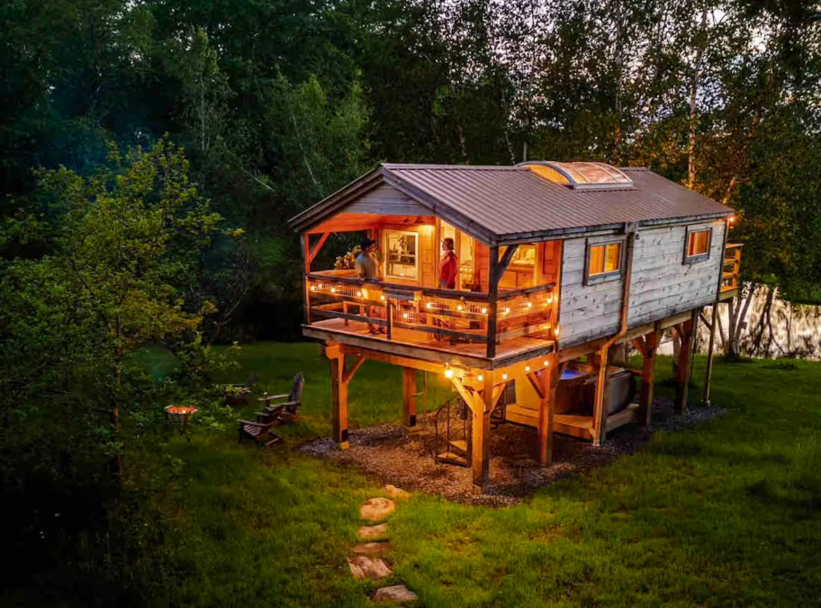Wooden glamping cabin on stilts with porch string lights, surrounded by trees, grass, chairs, and a fire pit in the yard.