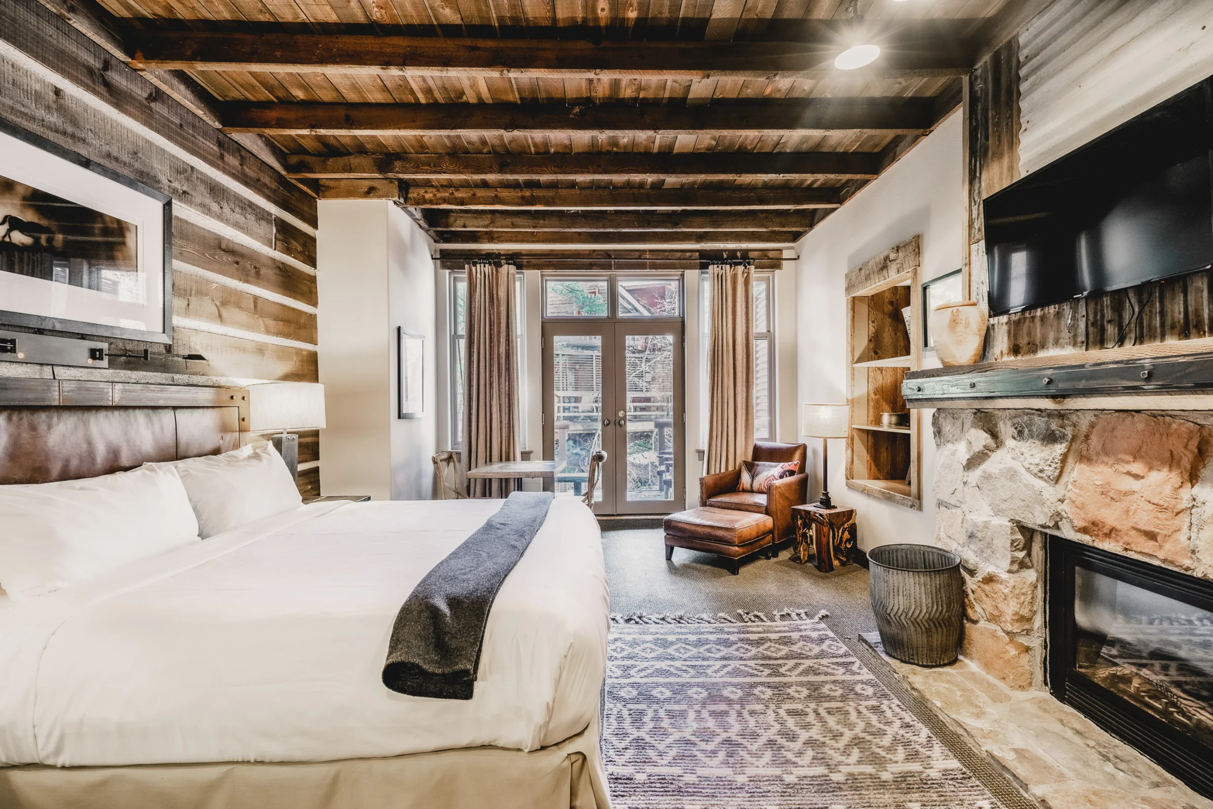 Rustic bedroom featuring a king bed, beamed ceiling, stone fireplace, leather chair, rug, and balcony doors.