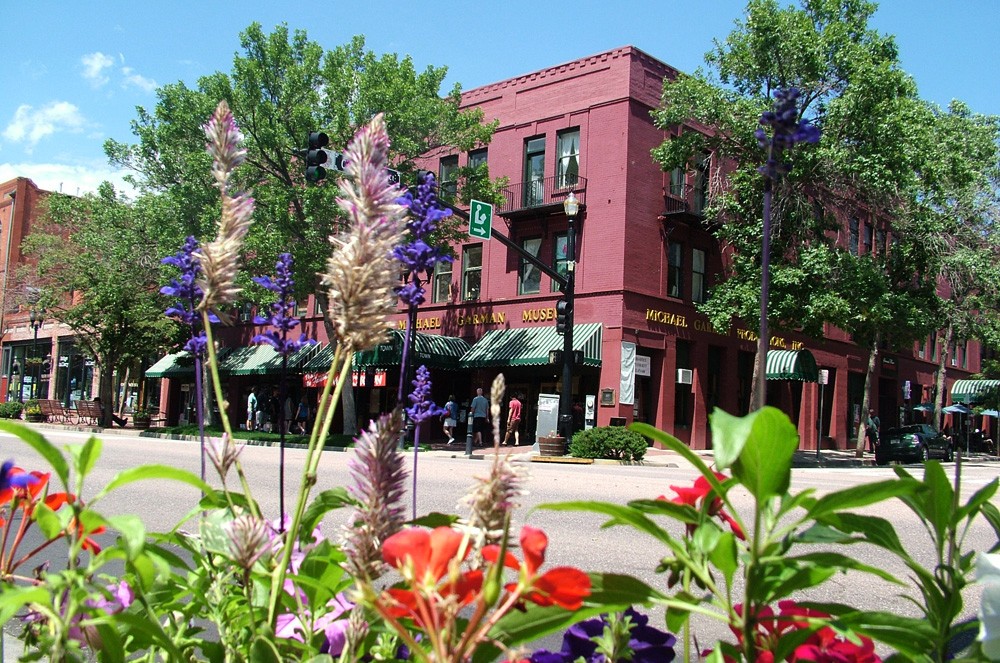 Red brick building with green awnings, trees, and colorful flowers in front on a sunny day.