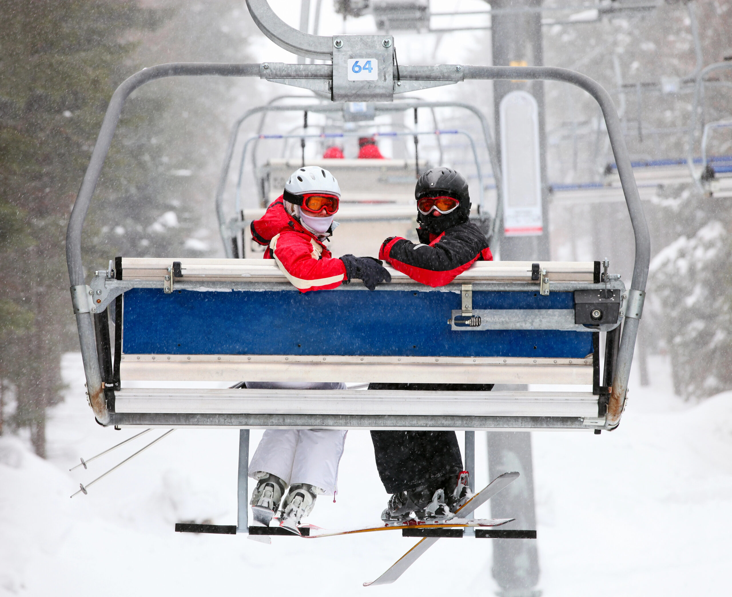 Two people in winter gear sit on a ski lift with ski poles, ascending a snowy mountain as snow falls around them.