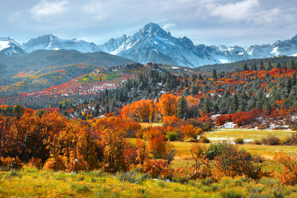 Snow-capped mountains tower over autumn trees and rolling hills beneath a partly cloudy sky.