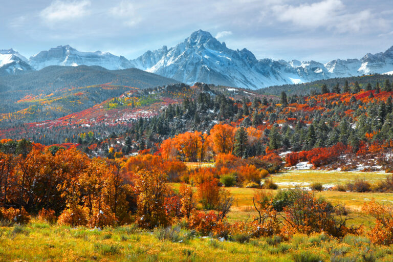 Snow-capped mountains tower over autumn trees and rolling hills beneath a partly cloudy sky.