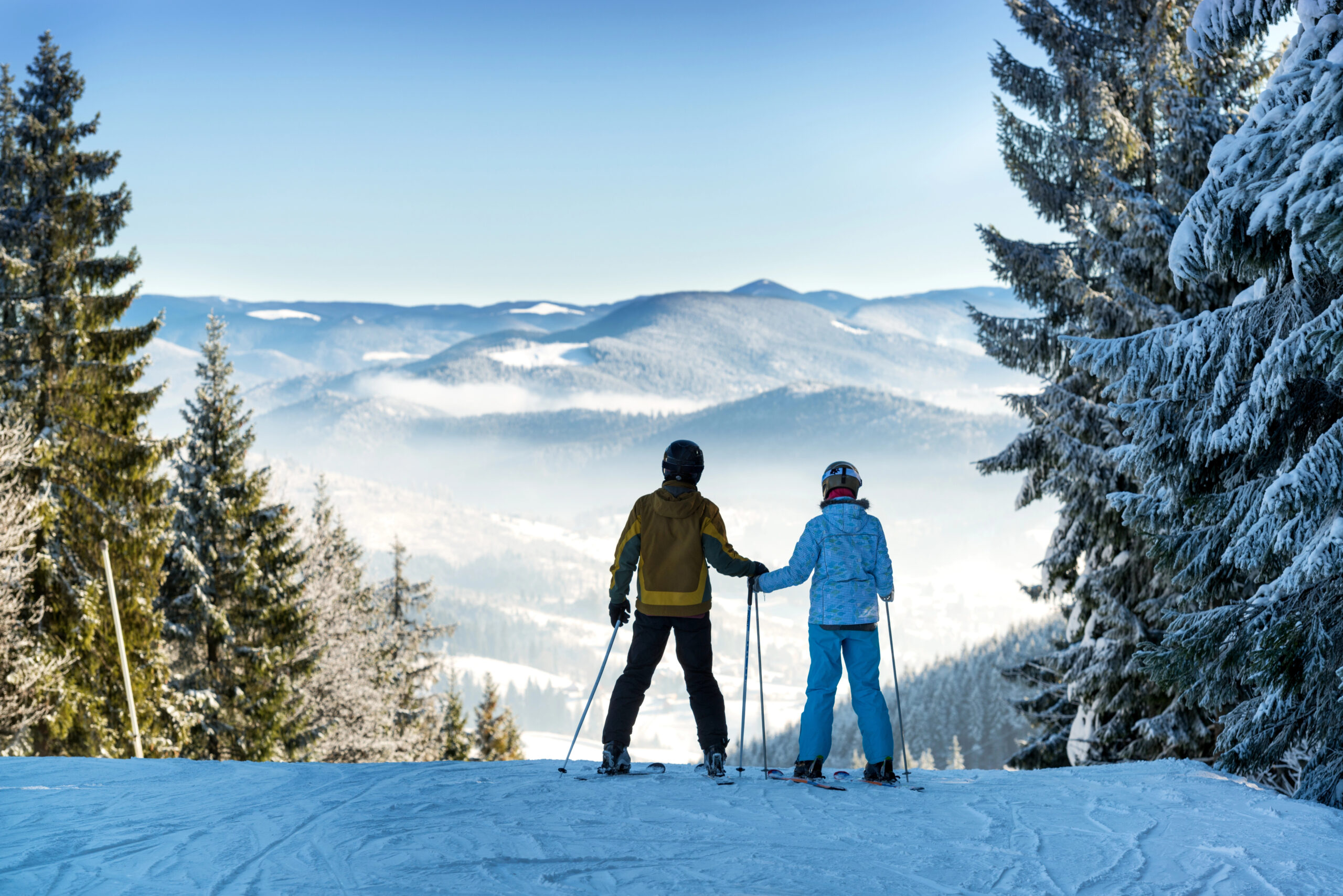A couple in ski gear holding hands at the top of a snow covered mountain slope.