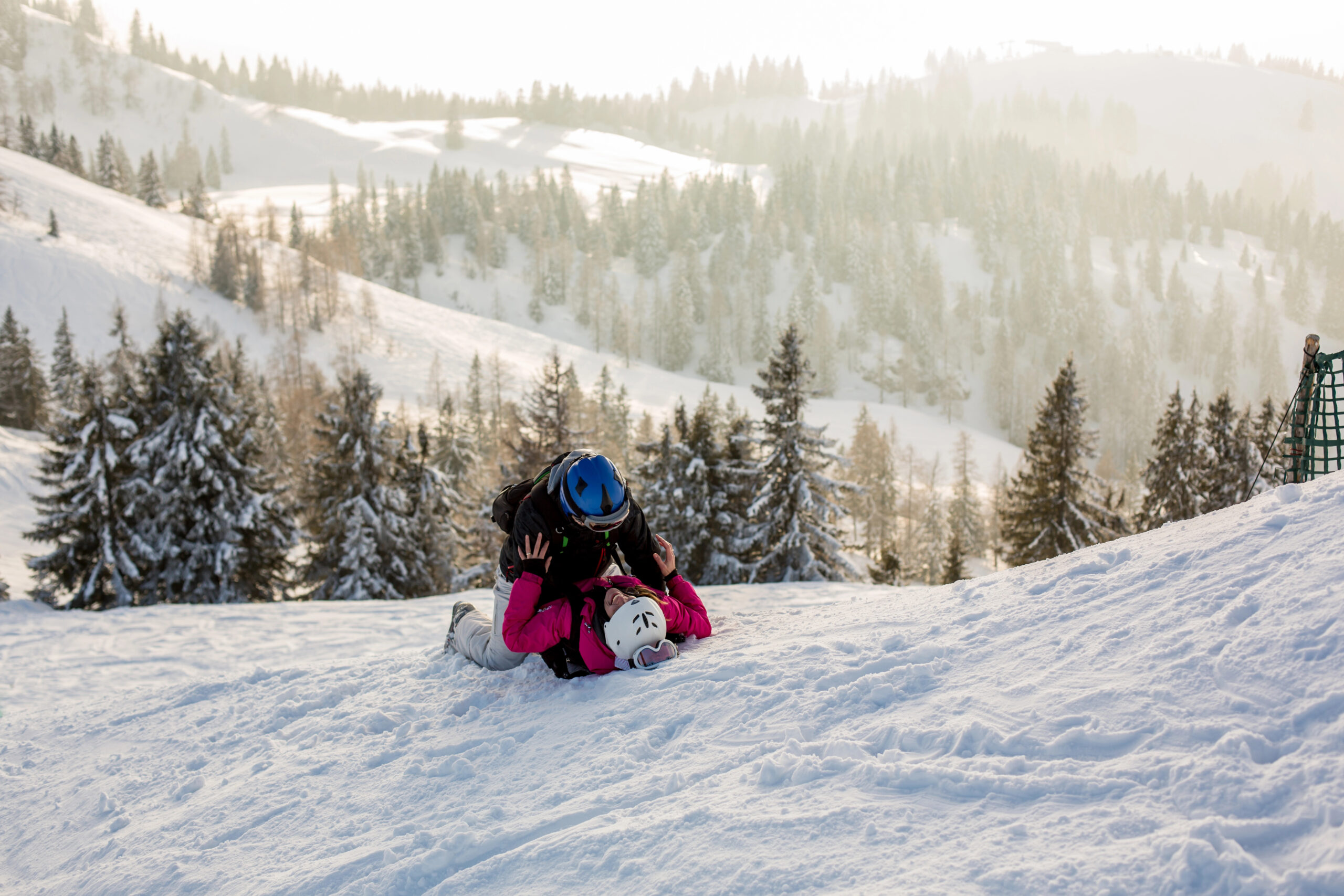 Two people in ski gear lie on a snowy mountain slope, one positioned on top of the other.