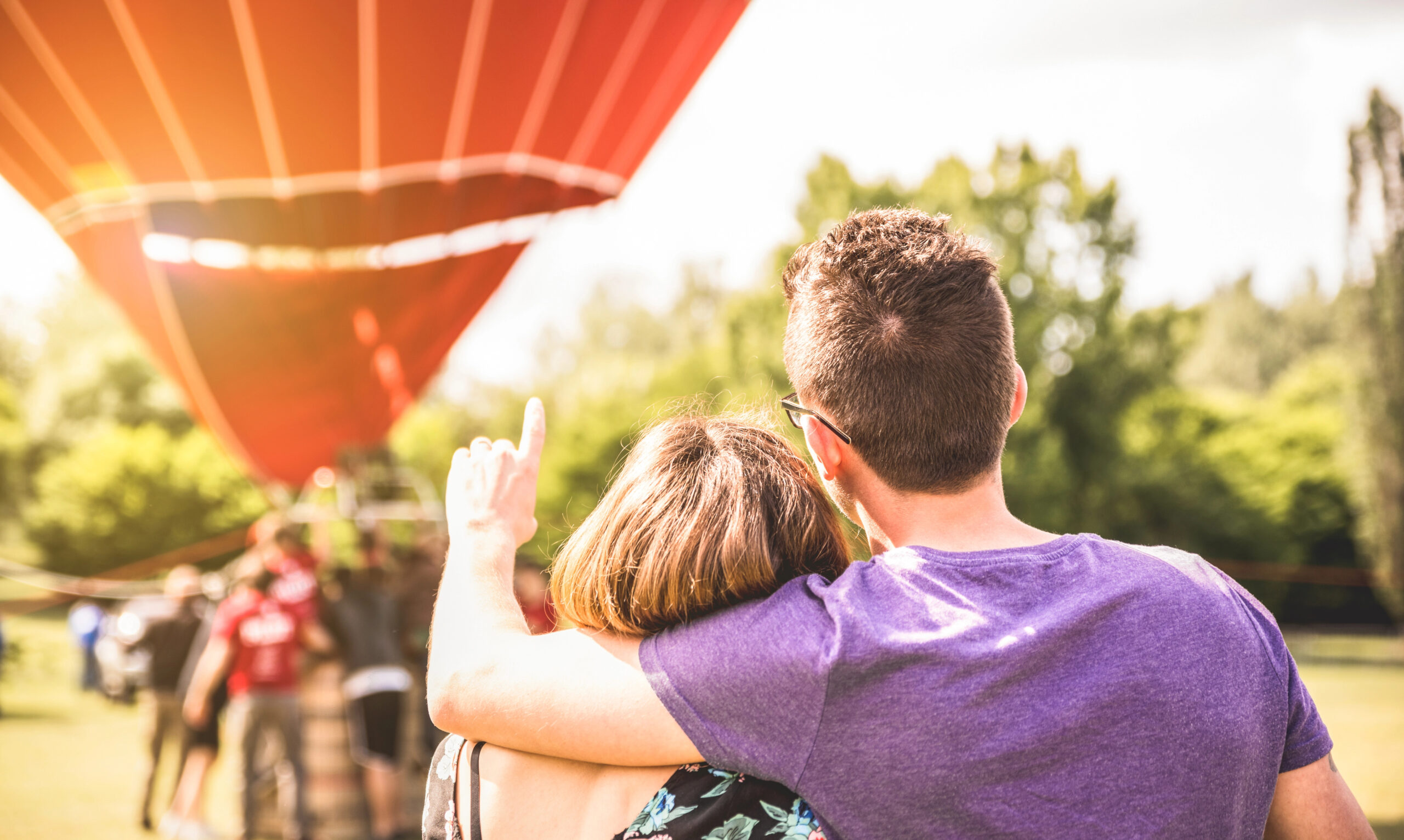 Two people embrace outdoors, pointing at a red hot air balloon ready to launch in a grassy, tree-lined Colorado Springs field.