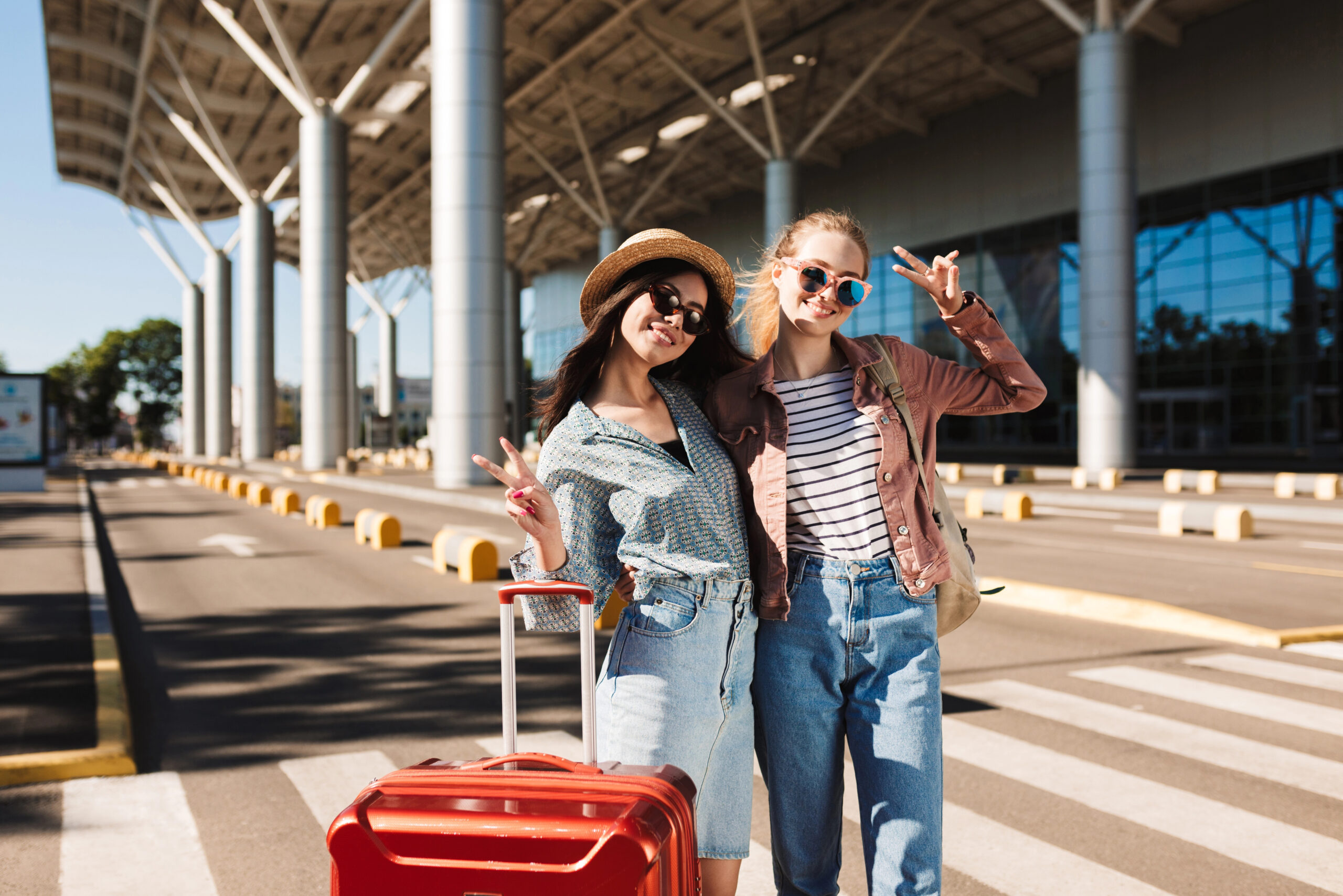 Two women with luggage smile and flash peace signs on an airport crosswalk, ready for their trip.