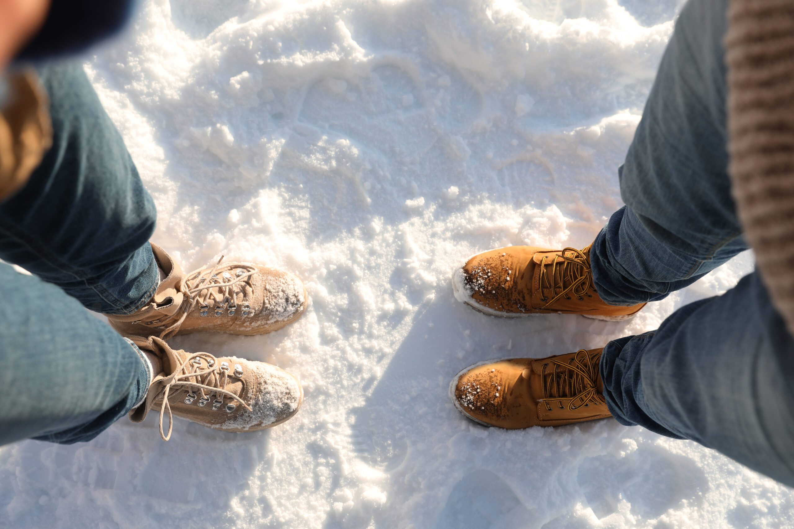 Two people in jeans and boots face each other on snowy ground, with snow on their boots.