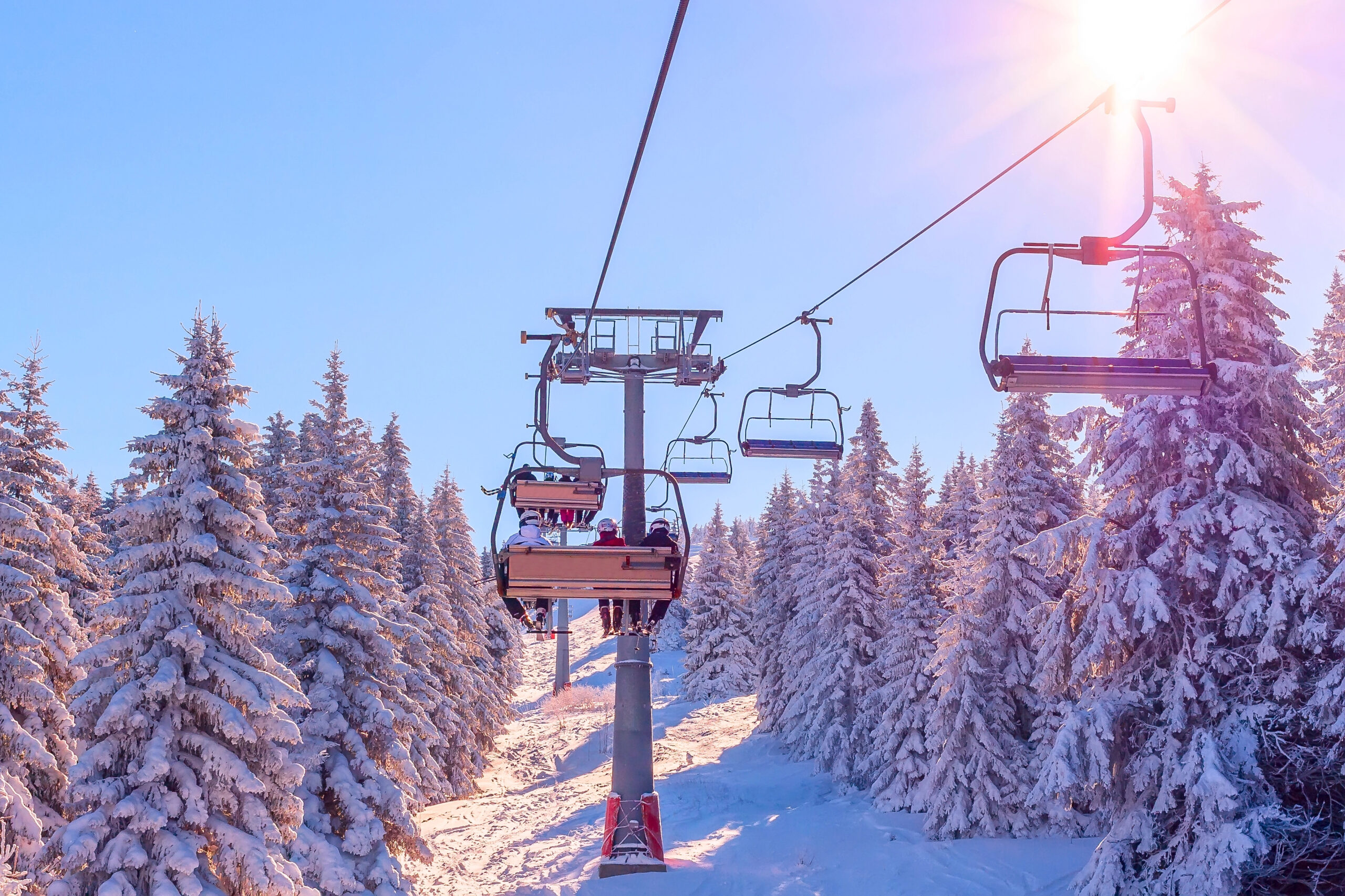 A ski lift transports people above a snowy slope bordered by pine trees under a sunny, clear blue sky.