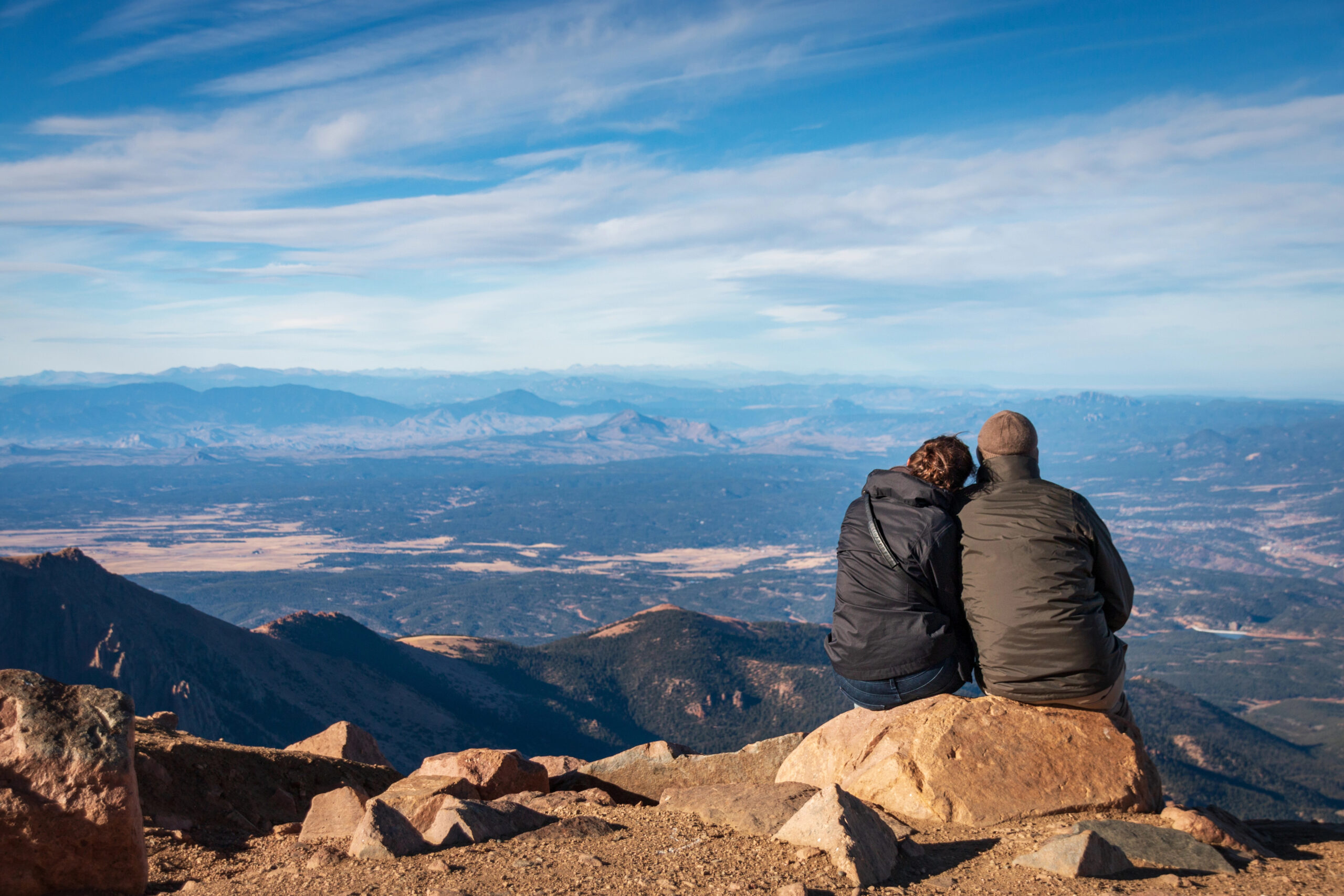 Two people sit on rocks overlooking a mountainous landscape, one resting their head on the other’s shoulder under a cloudy sky.
