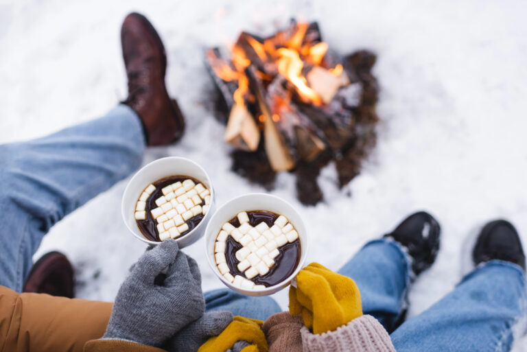 Two people in winter clothes and gloves sit on snow by a campfire, holding mugs of hot chocolate with marshmallows.