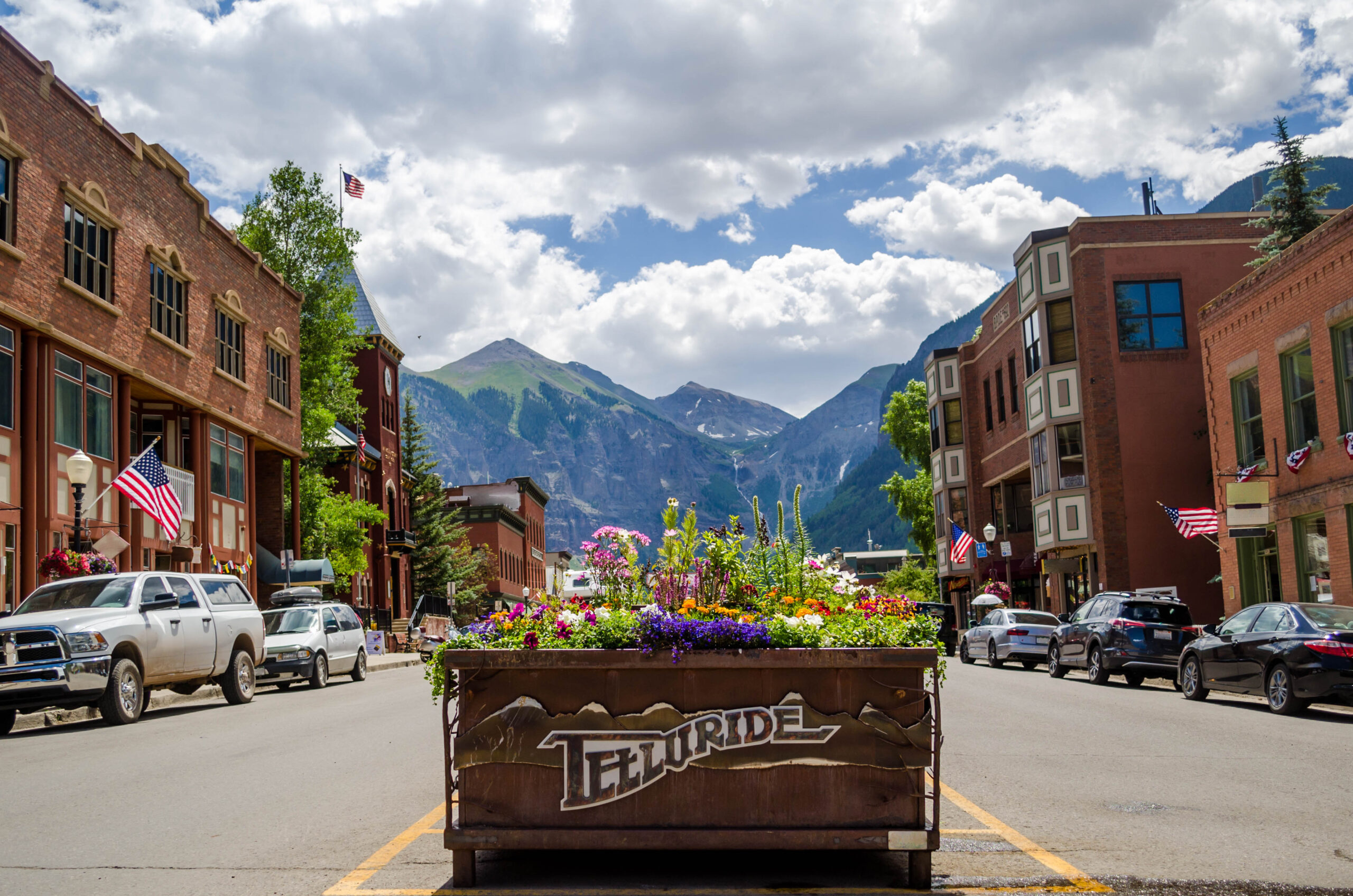 Street in Telluride, Colorado with central flower planter, historic buildings, parked cars, and mountains beneath a partly cloudy sky. It's one of the best romantic mountain getaways in Colorado!