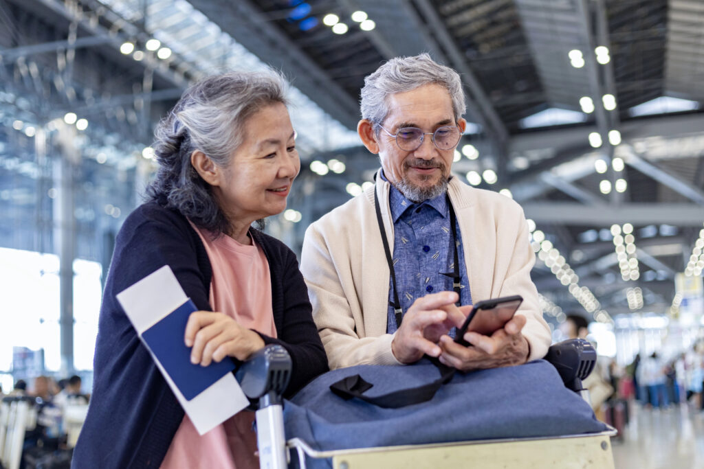 An older couple in an airport terminal, one holding travel documents and the other using a smartphone, with luggage in front.