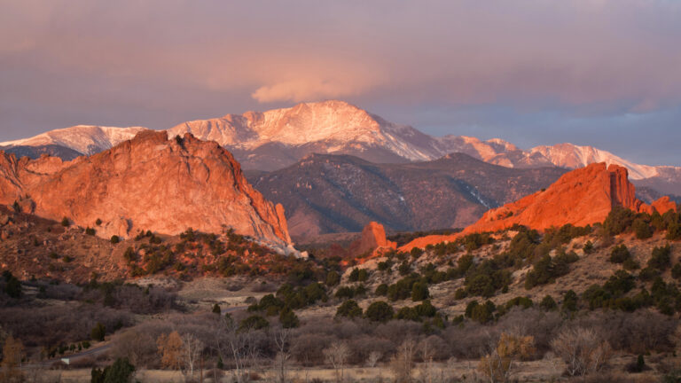 Red rock formations and scattered trees lie before a snow-capped mountain beneath a partly cloudy sky.