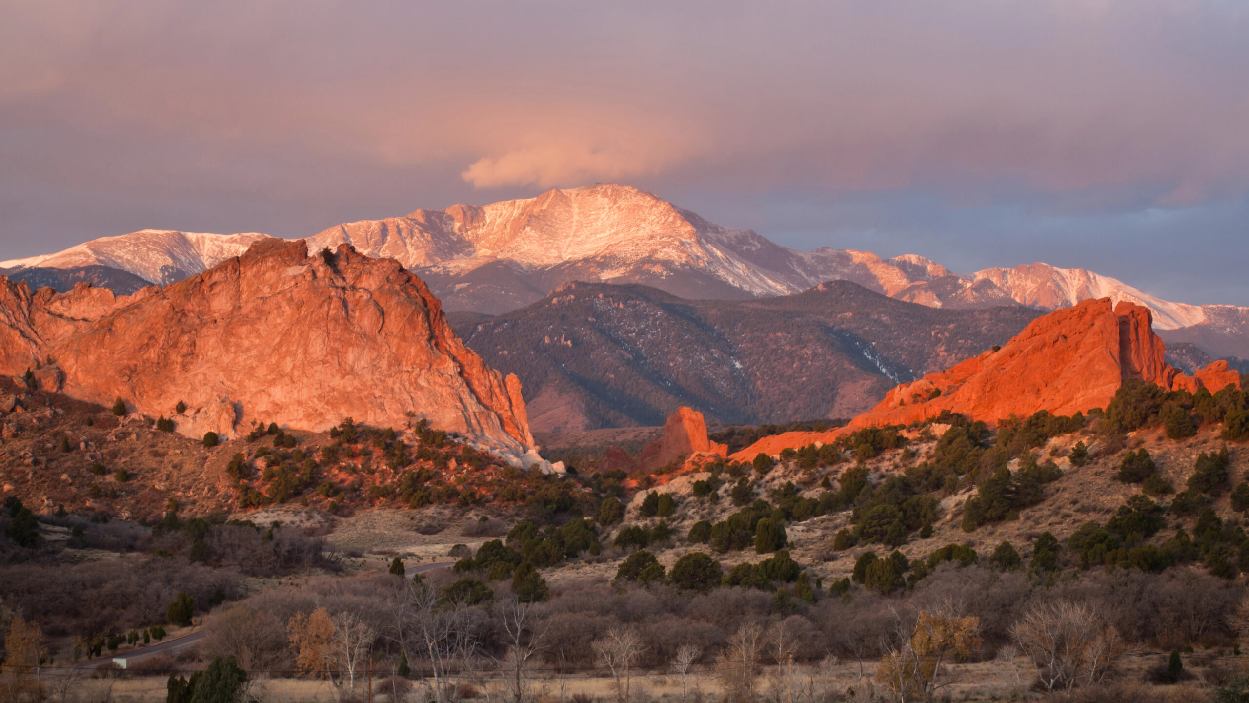 Red rock formations and scattered trees lie before a snow-capped mountain beneath a partly cloudy sky.