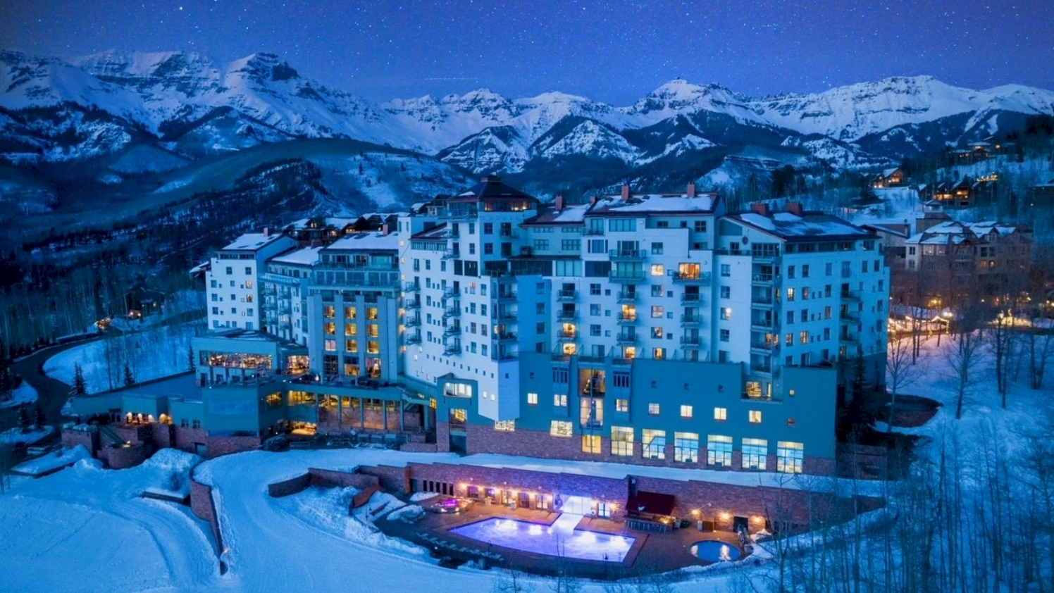 The Peaks Resort & Spa with glowing outdoor pools, snowy forest, and mountain backdrop beneath a starry night sky. It's one of the most romantic ski resorts in the USA!