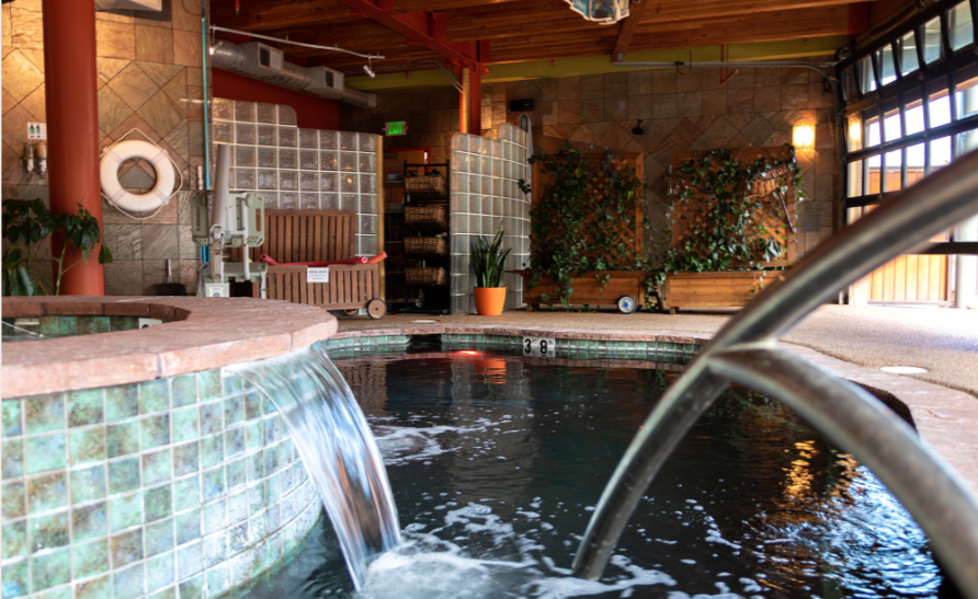 Indoor pool with waterfall, tiled walls, potted plants, benches, and sunlit glass doors in Colorado Springs.
