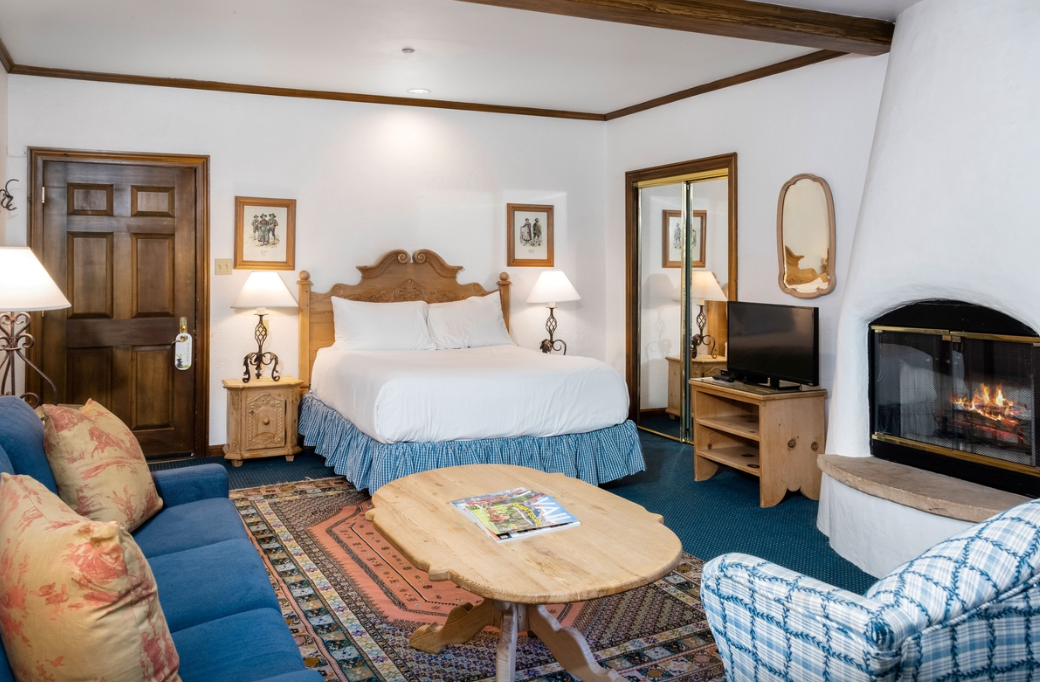 A cozy hotel room features a bed, sofa, wooden table, TV, fireplace, lamps, and framed wall art.